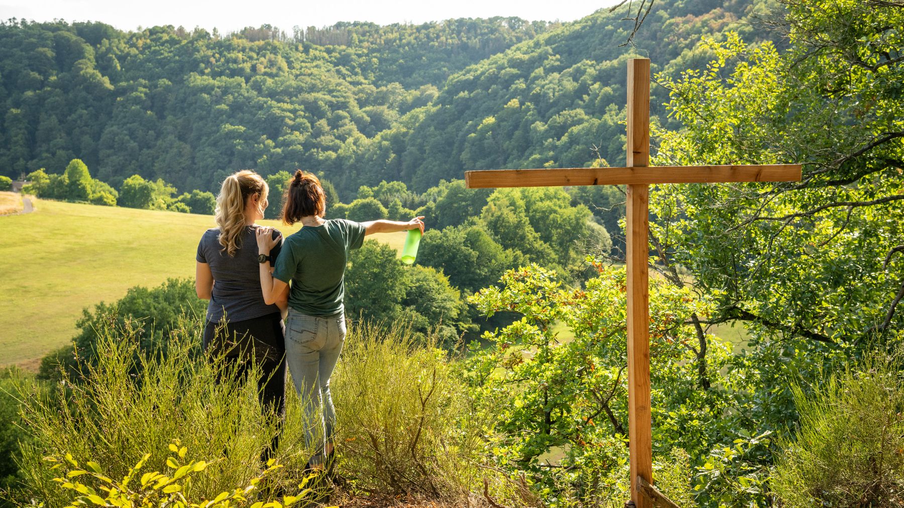 Zwei Frauen stehen auf einer Bergkuppe in der Nähe eines großen Holzkreuzes und blicken auf ein malerisches, bewaldetes Tal. Eine von ihnen zeigt mit einer Karte voraus, was darauf hindeutet, dass sie gemeinsam wandern oder die Gegend erkunden.